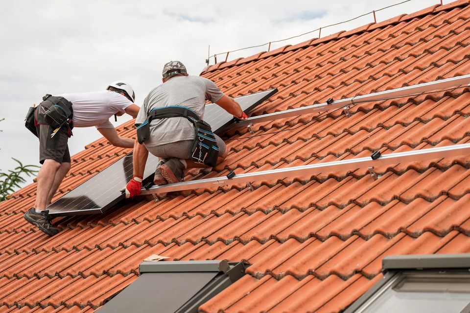 Deux techniciens en combinaisons vertes et casques blancs installent des panneaux photovoltaïques sur une toiture résidentielle en tuiles sous un ciel bleu dégagé, environnement verdoyant visible en arrière-plan