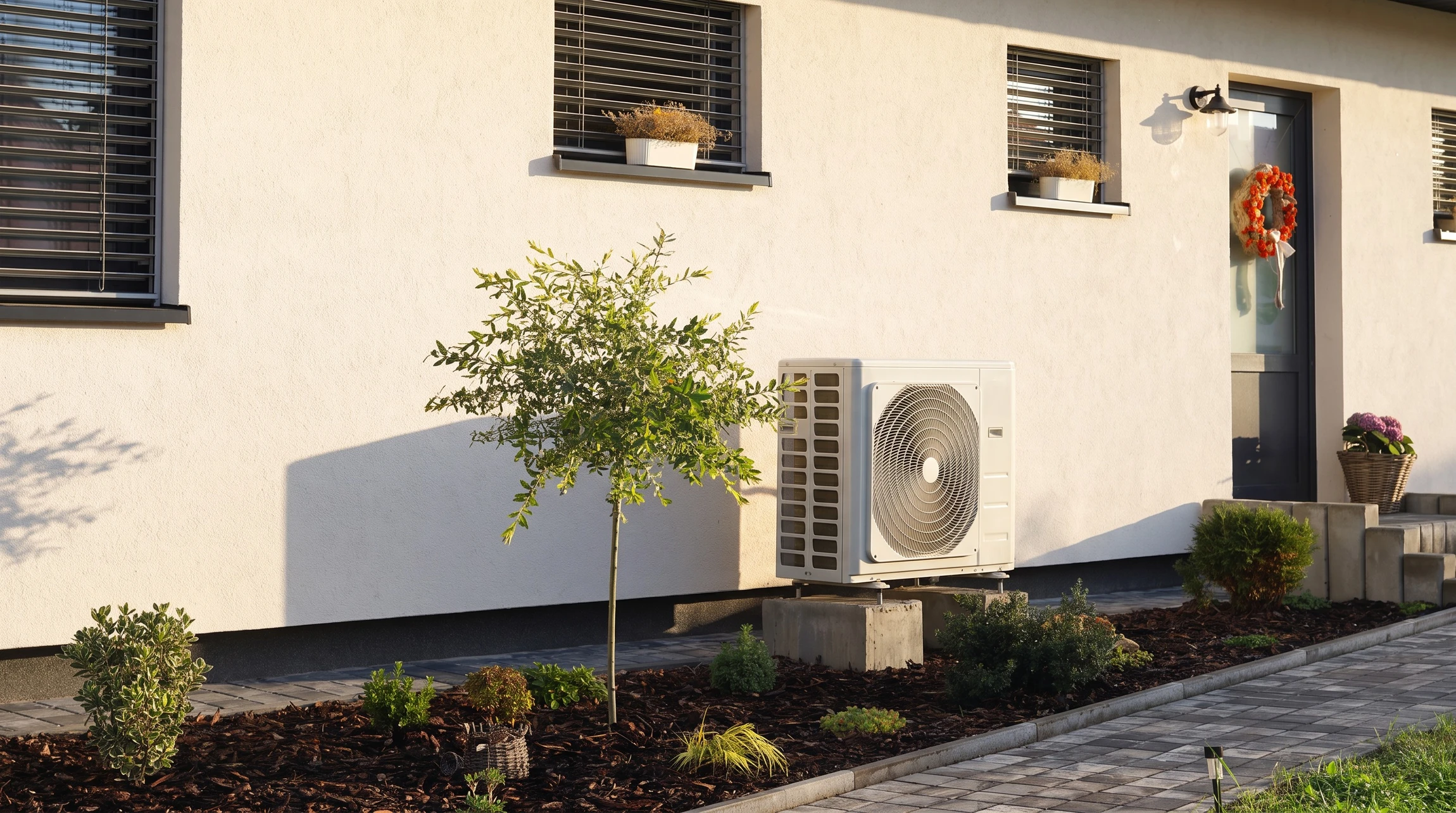 Photographie en plein jour d'une unité extérieure de pompe à chaleur air-air récemment installée sur la façade d'une maison résidentielle, montrant clairement le boîtier blanc ou gris avec grille de ventilation. La résidence adjacente doit être visible en arrière-plan, avec un jardin ou cour bien entretenue. L'image doit transmettre la modernité, la fiabilité et l'intégration harmonieuse du système dans un habitat contemporain.